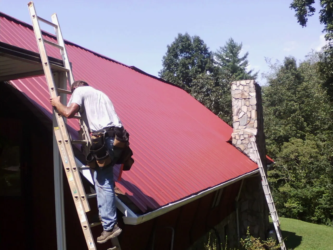 Roofer on a ladder beside a red metal roof and stone chimney — Soffit & Fascia Repair by Roofing Brothers in Surgoinsville